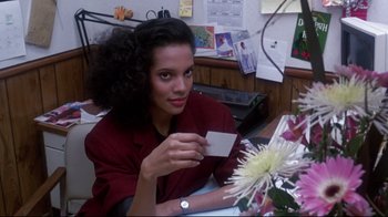 Movie still from “Coming to America” (1988), directed by John Landis – A woman sitting at a desk holding a business card; Close Up shot, Over the shoulder angle