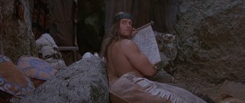 Movie still from “Conan the Barbarian” (1982), directed by John Milius – A man in native american garb sitting on top of a tree; Medium shot, Low angle