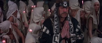 Movie still from “Conan the Barbarian” (1982), directed by John Milius – A man with a long beard wearing a headdress and praying in front of a group of people with lit candles; Medium shot, Low angle