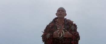 Movie still from “Conan the Barbarian” (1982), directed by John Milius – A bald man wearing a necklace and an animal skin cape; Medium shot, Low angle