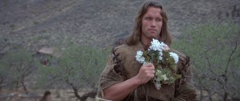 Movie still from “Conan the Barbarian” (1982), directed by John Milius – A man holding flowers in his hand while standing in a field; Medium shot, Over the shoulder angle