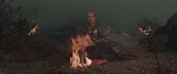 Movie still from “Conan the Barbarian” (1982), directed by John Milius – A man sitting on the ground next to a fire; Wide shot, Low angle
