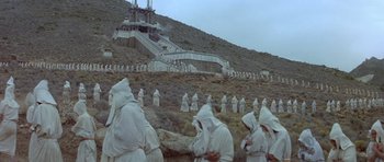 Movie still from “Conan the Barbarian” (1982), directed by John Milius – A group of people dressed in white standing in front of a mountain; Extreme Wide shot, High angle
