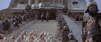 Movie still from “Conan the Barbarian” (1982), directed by John Milius – A group of people dressed in white and black are standing in front of a building; Wide shot, High angle