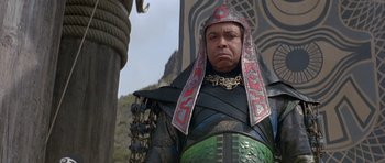 Movie still from “Conan the Barbarian” (1982), directed by John Milius – A man in a costume is standing next to a pole; Medium shot, Low angle