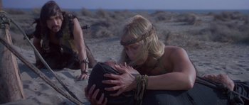 Movie still from “Conan the Barbarian” (1982), directed by John Milius – A man and a woman kissing on the beach; Medium shot, Over the shoulder angle