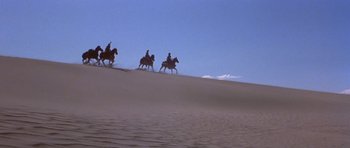 Movie still from “Conan the Barbarian” (1982), directed by John Milius – A group of people riding horses across a sand dune; Extreme Wide shot, Low angle