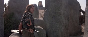 Movie still from “Conan the Barbarian” (1982), directed by John Milius – A man holding a sword standing in front of a stone wall; Medium shot, Over the shoulder angle