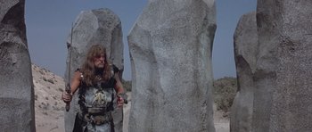 Movie still from “Conan the Barbarian” (1982), directed by John Milius – A man standing next to two large rocks; Wide shot, Low angle