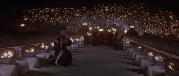 Movie still from “Conan the Barbarian” (1982), directed by John Milius – A group of people in native american garb are sitting in front of a group of lights; Extreme Wide shot, High angle