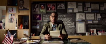 Movie still from “Da 5 Bloods” (2020), directed by Spike Lee – A man sitting in front of a chalkboard holding a piece of paper; Medium shot, Over the shoulder angle