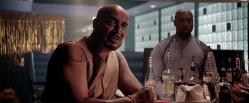 Movie still from “Debt Collectors” (2020), directed by Jesse V. Johnson – A man sitting at a table with a bottle of alcohol; Medium shot, Over the shoulder angle