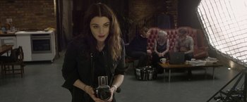 Movie still from “Disobedience” (2017), directed by Sebastián Lelio – A woman holding a camera in a room with two people; Medium shot, Over the shoulder angle
