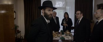Movie still from “Disobedience” (2017), directed by Sebastián Lelio – A man in a hat and glasses standing in front of a table; Medium shot, Over the shoulder angle