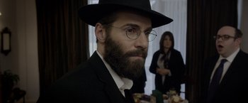Movie still from “Disobedience” (2017), directed by Sebastián Lelio – A man with a beard wearing glasses and a hat; Close Up shot, Over the shoulder angle