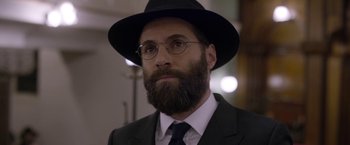 Movie still from “Disobedience” (2017), directed by Sebastián Lelio – A man in a suit and hat with a beard; Close Up shot, Over the shoulder angle