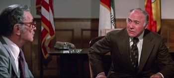 Movie still from “Earthquake” (1974), directed by Mark Robson – A man sitting in front of a desk in front of flags; Medium shot, Over the shoulder angle