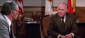 Movie still from “Earthquake” (1974), directed by Mark Robson – An older man in a suit sitting in a chair in front of an american flag; Close Up shot, Over the shoulder angle
