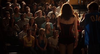 Movie still from “Easy A” (2010), directed by Will Gluck – A group of people sitting in a room watching a woman; Wide shot, Over the shoulder angle
