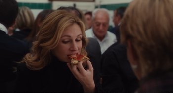 Movie still from “Eat Pray Love” (2010), directed by Ryan Murphy – A woman eating a piece of pizza in front of a group of people; Close Up shot, Over the shoulder angle