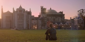 Movie still from “Fate: The Winx Saga” (2021), created by Brian Young – A man sitting on the grass in front of a building; Extreme Wide shot, Over the shoulder angle