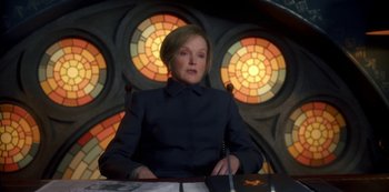 Movie still from “Fate: The Winx Saga” (2021), created by Brian Young – A woman sitting at a table in front of stained glass windows; Medium shot, Low angle