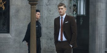 Movie still from “Fate: The Winx Saga” (2021), created by Brian Young – A man in a suit and tie standing next to a pole; Medium shot, Over the shoulder angle