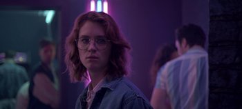 Movie still from “Black Mirror” (2011), created by Charlie Brooker – A woman with red hair and glasses in a purple room; Medium shot, Over the shoulder angle