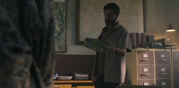 Movie still from “Feria: The Darkest Light” (2022), created by Carlos Montero – A man standing in front of a wall holding a book; Medium shot, Over the shoulder angle