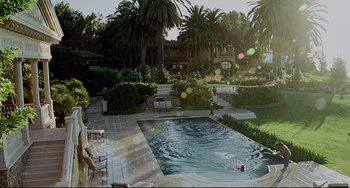 Movie still from “Funny People” (2009), directed by Judd Apatow – A man swimming in a pool surrounded by chairs and trees; Extreme Wide shot, High angle