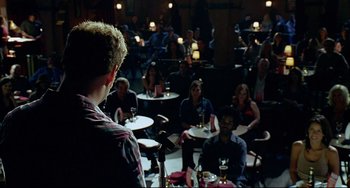 Movie still from “Funny People” (2009), directed by Judd Apatow – A group of people sitting at tables in front of a microphone; Wide shot, Over the shoulder angle