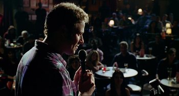 Movie still from “Funny People” (2009), directed by Judd Apatow – A man holding a microphone while standing in front of a group of people; Medium shot, Over the shoulder angle
