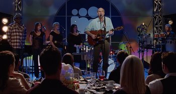 Movie still from “Funny People” (2009), directed by Judd Apatow – A group of people sitting around a table with a man playing a guitar; Wide shot, Over the shoulder angle