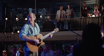 Movie still from “Funny People” (2009), directed by Judd Apatow – An older man playing a guitar in front of a crowd of onlookers; Medium shot, Over the shoulder angle