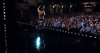 Movie still from “Funny People” (2009), directed by Judd Apatow – A man riding a bike in front of an audience; Extreme Wide shot, High angle