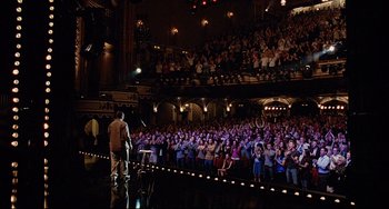 Movie still from “Funny People” (2009), directed by Judd Apatow – A crowd of people watching a man sing on a stage; Extreme Wide shot, High angle