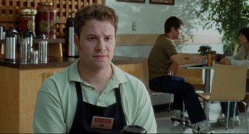 Movie still from “Funny People” (2009), directed by Judd Apatow – A man in an apron sitting at a table with a cup of coffee in front of him; Medium shot, Over the shoulder angle