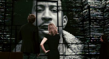 Movie still from “Gamer” (2009), directed by Mark Neveldine – A man and a woman standing in front of a picture of a man; Medium shot, Over the shoulder angle