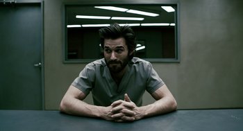 Movie still from “Gamer” (2009), directed by Mark Neveldine – A man sitting at a table with his arms crossed; Medium shot, Low angle