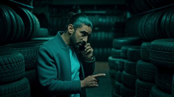 Movie still from “Ganglands” (2021), created by Julien Leclercq – A man with blue hair talking on a cell phone; Medium shot, Over the shoulder angle