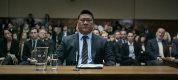 Movie still from “Black Mirror” (2011), created by Charlie Brooker – A man sitting at a table in front of an audience; Medium shot, Low angle