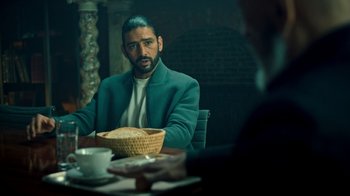 Movie still from “Ganglands” (2021), created by Julien Leclercq – A man sitting at a table with a bowl of bread; Medium shot, Over the shoulder angle