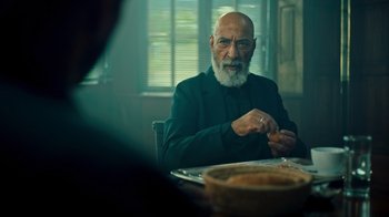 Movie still from “Ganglands” (2021), created by Julien Leclercq – An older man sitting at a table in front of a bowl of food; Medium shot, Over the shoulder angle