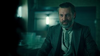 Movie still from “Ganglands” (2021), created by Julien Leclercq – A man with a beard wearing a suit and tie; Medium shot, Low angle