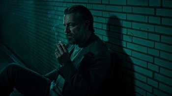 Movie still from “Ganglands” (2021), created by Julien Leclercq – A man smoking a cigarette while leaning against a brick wall; Medium shot, Low angle