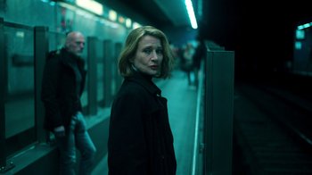 Movie still from “Ganglands” (2021), created by Julien Leclercq – A woman standing on a train platform next to a wall; Medium shot, Low angle