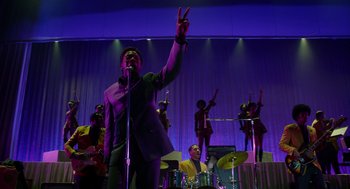 Movie still from “Get on Up” (2014), directed by Tate Taylor – A man holding a peace sign while standing next to a microphone; Medium shot, Low angle