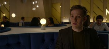 Movie still from “Black Mirror” (2011), created by Charlie Brooker – A man sitting in front of a table in a room; Medium shot, Over the shoulder angle