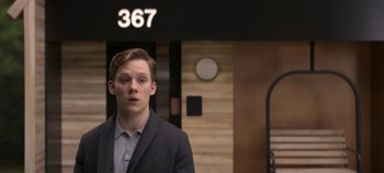 Movie still from “Black Mirror” (2011), created by Charlie Brooker – A man in a suit standing in front of a 3 6 7 sign; Close Up shot, Low angle