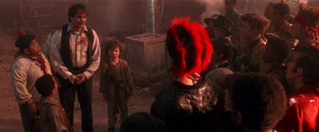 Movie still from “Hook” (1991), directed by Steven Spielberg – A person with a red feather headdress is standing next to a crowd; Medium shot, Over the shoulder angle